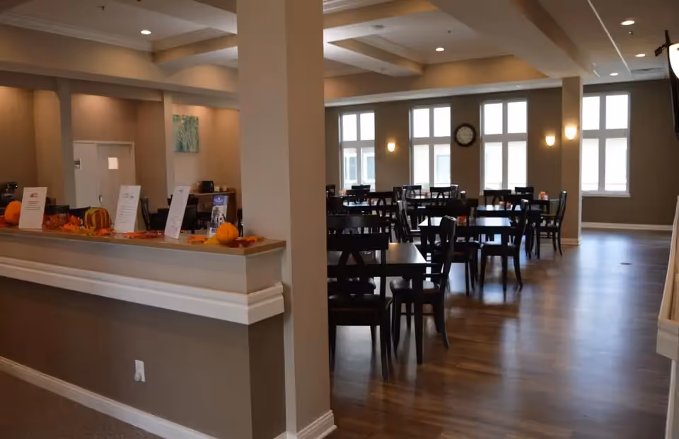 Interior view of a dining room in an assisted living facility with multiple dark wooden tables and chairs arranged neatly on a wooden floor. The room has large windows allowing natural light, beige walls, and a clock mounted on the wall. A counter decorated with small pumpkins and autumn leaves is visible in the foreground.