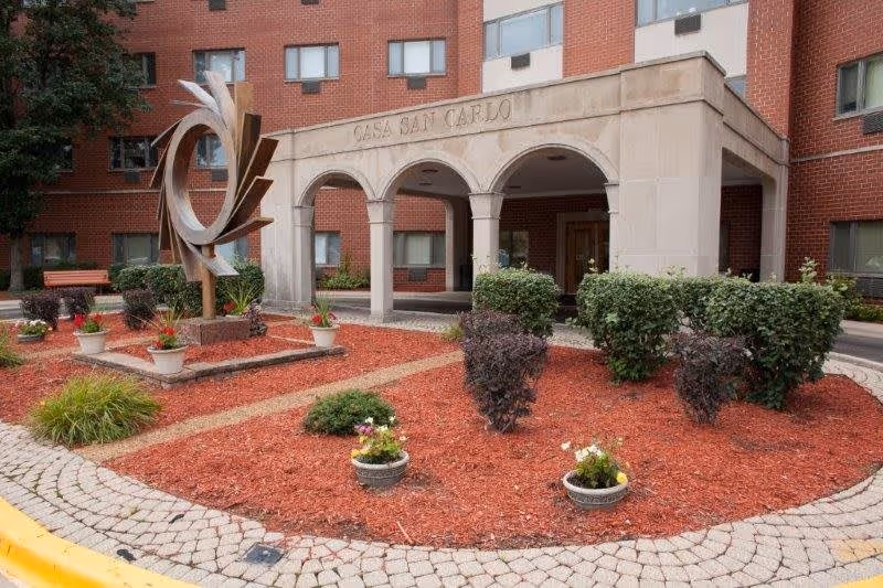 Exterior view of a senior living facility named Casa San Carlo featuring a brick building with multiple windows, an entrance with three arches, landscaped garden beds with red mulch, shrubs, potted flowers, and a modern metal sculpture.