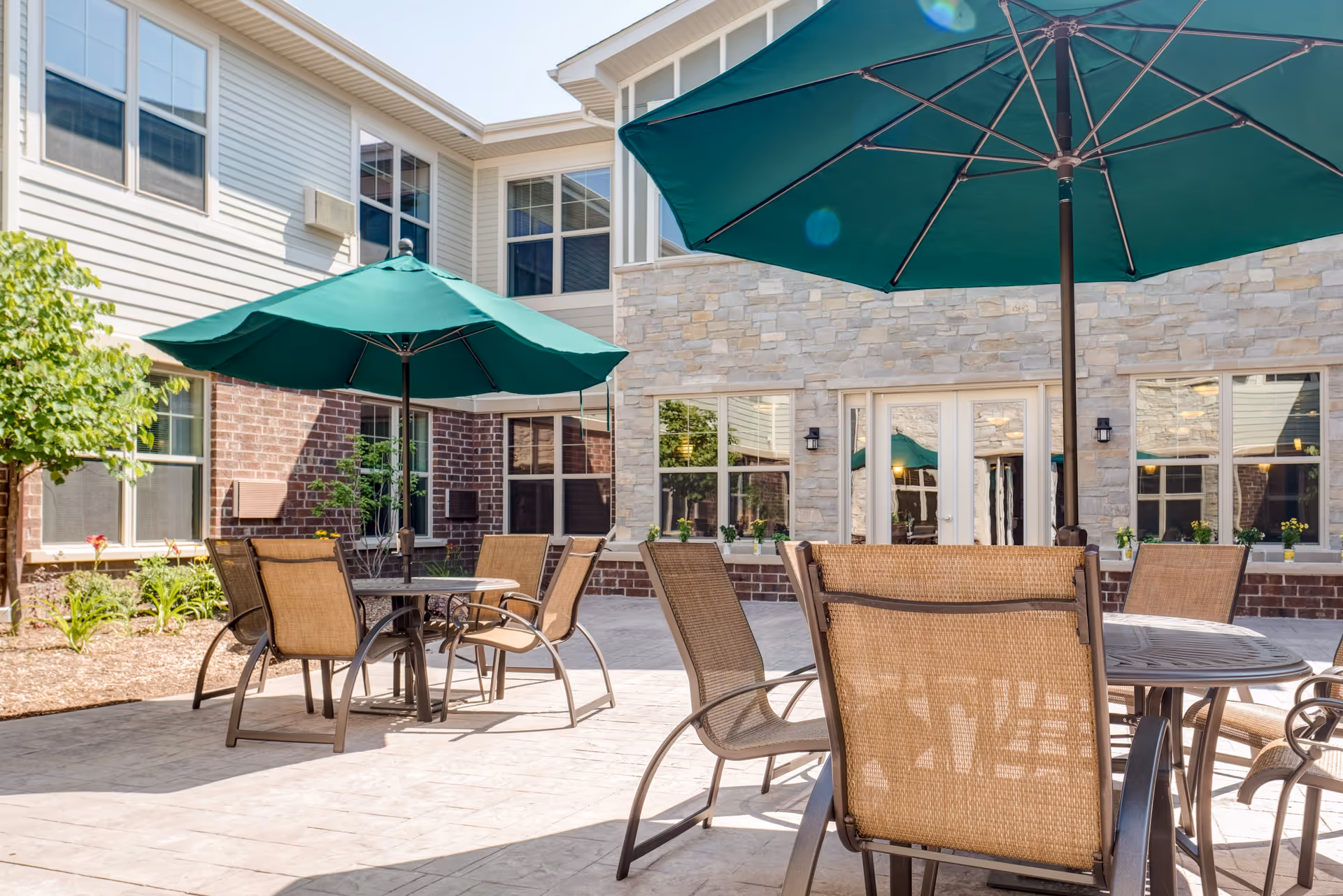 Outdoor patio area at Heritage Lake Country Senior Living with several round tables and chairs under large green umbrellas, surrounded by a building with brick and stone exterior walls and multiple windows.