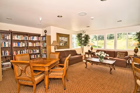 A cozy living room area with a brown carpet, two brown sofas, a wooden coffee table with a vase of white flowers, a wooden table with four upholstered chairs, a bookshelf filled with books, a floor lamp, a framed painting on the wall, and large windows showing greenery outside.