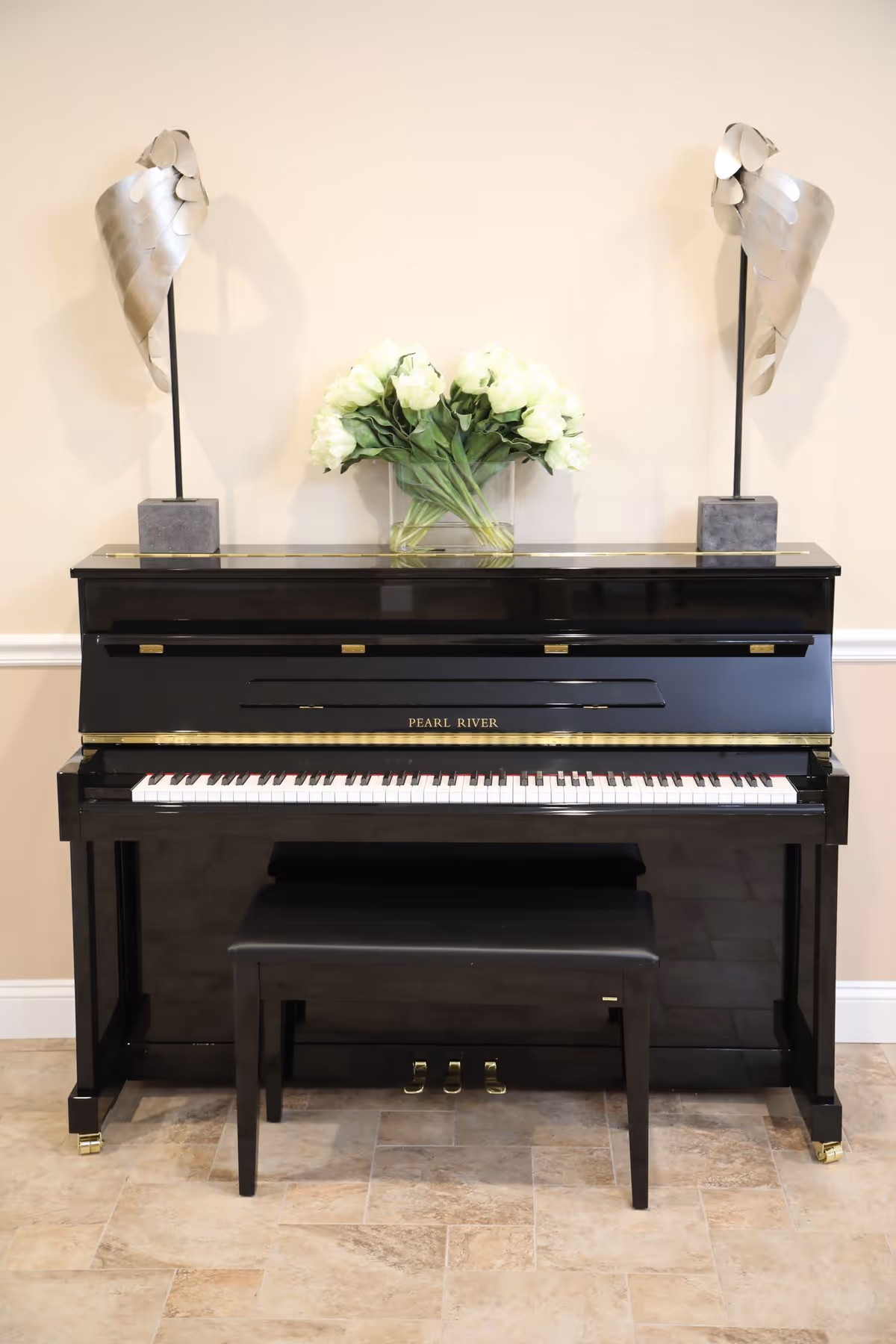 A black upright Pearl River piano with a matching black bench in front of a beige wall. On top of the piano, there is a clear vase with white flowers and two modern silver sculptures on either side.