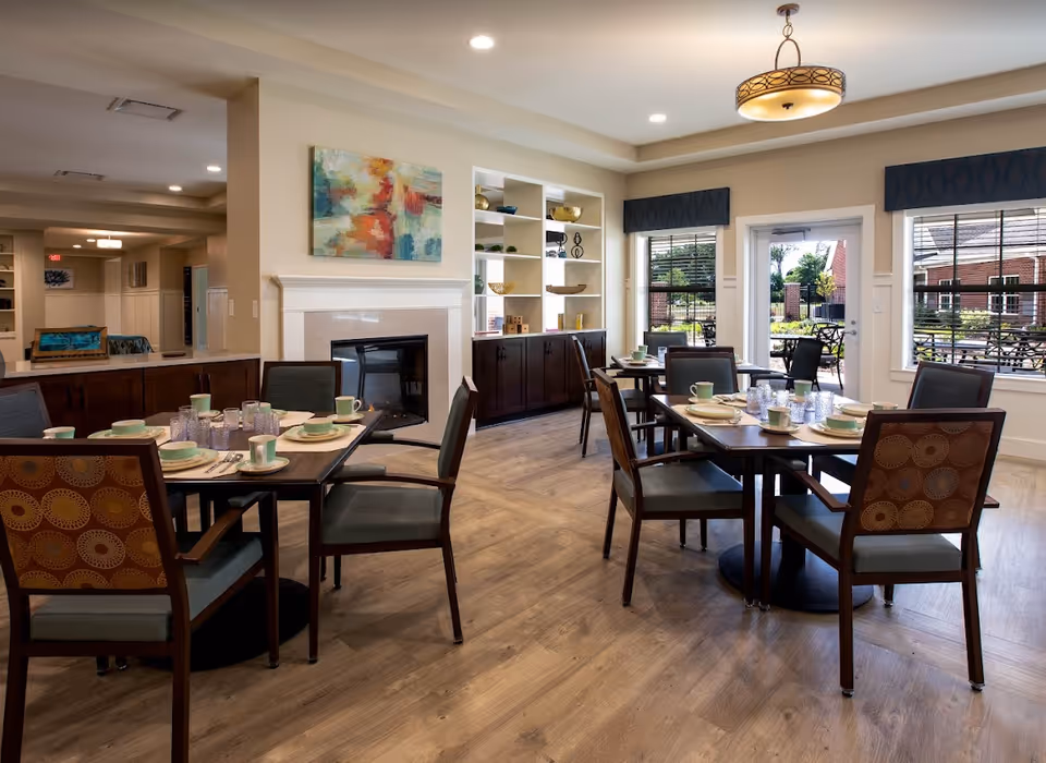 A well-lit dining room with wooden tables and chairs set with plates, cups, and glasses. The room features a fireplace with a colorful abstract painting above it, built-in shelves with decorative items, large windows with dark valances, and a door leading to an outdoor patio area.