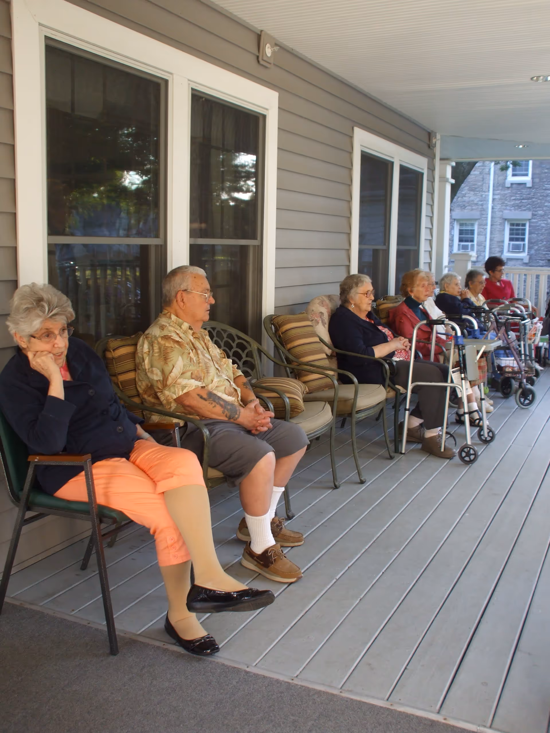 A group of elderly people sitting on chairs and benches on a covered porch outside a building. Some have walkers beside them, and the porch has gray wooden flooring and large windows along the wall.