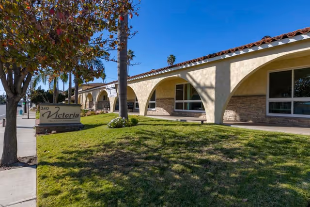 Front exterior of Victoria Healthcare and Rehabilitation featuring a low stucco building with arches, a lawn, palm trees, and a Victoria sign.