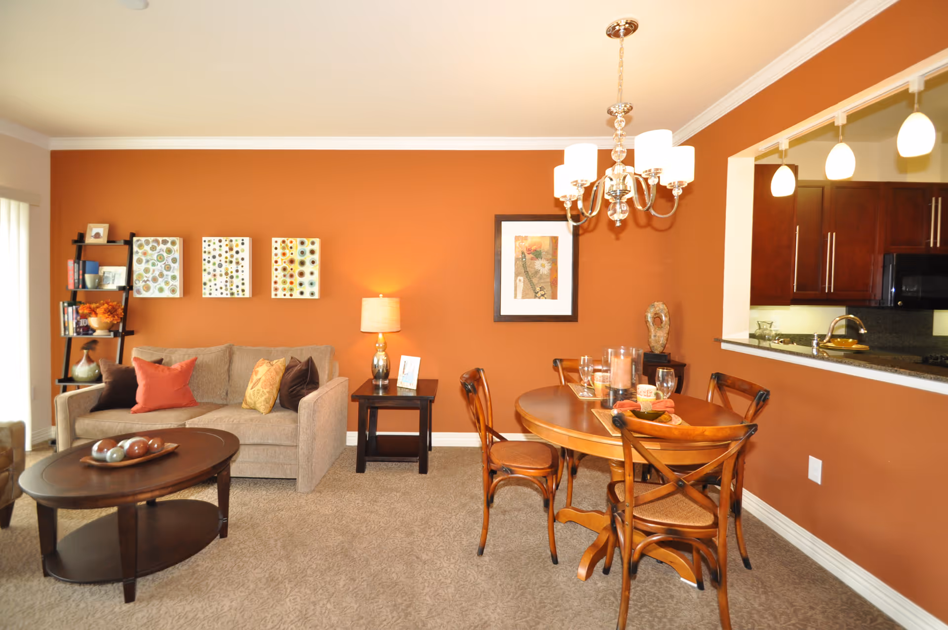 A cozy living and dining area with an orange accent wall. The living room features a beige sofa with colorful throw pillows, a dark wooden coffee table with decorative balls, and a black shelving unit with books and decor. The dining area has a wooden table with four chairs, a chandelier above, and a pass-through window to a kitchen with dark cabinets and pendant lights.