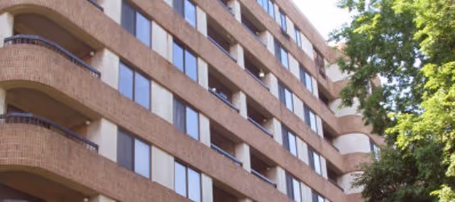 Multi-story brick building facade with repeating windows and curved balconies and a tree on the right.