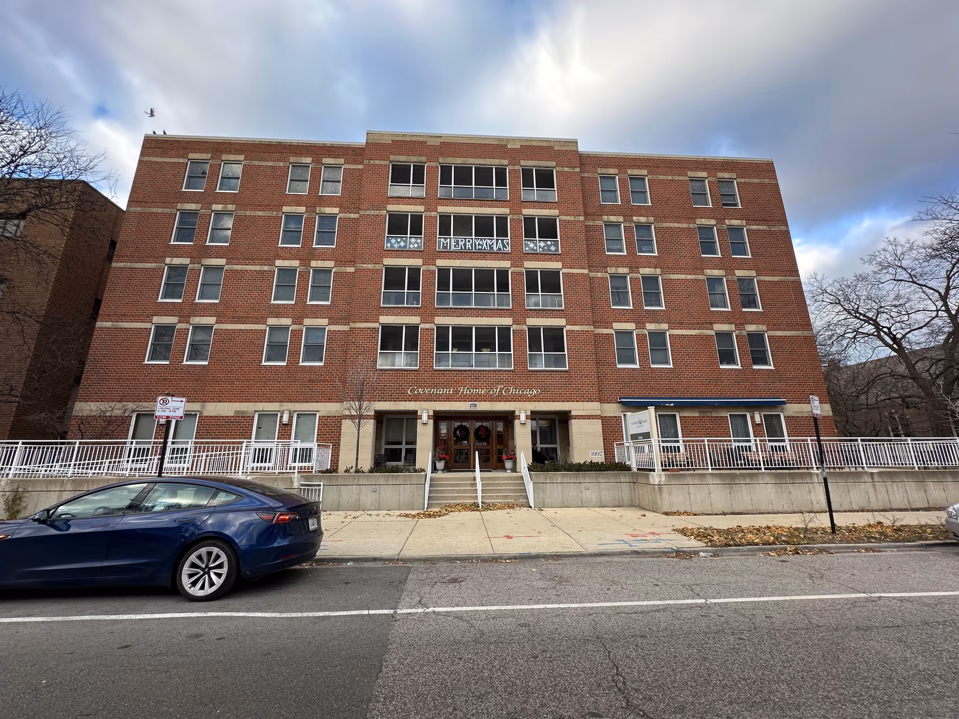 Front exterior view of a four-story brick building with multiple windows and a central entrance. The building has a sign above the entrance that reads 'Covenant Home of Chicago'. There are two wreaths on the entrance doors and a blue car parked on the street in front. The sky is cloudy and there are leafless trees on either side of the building.