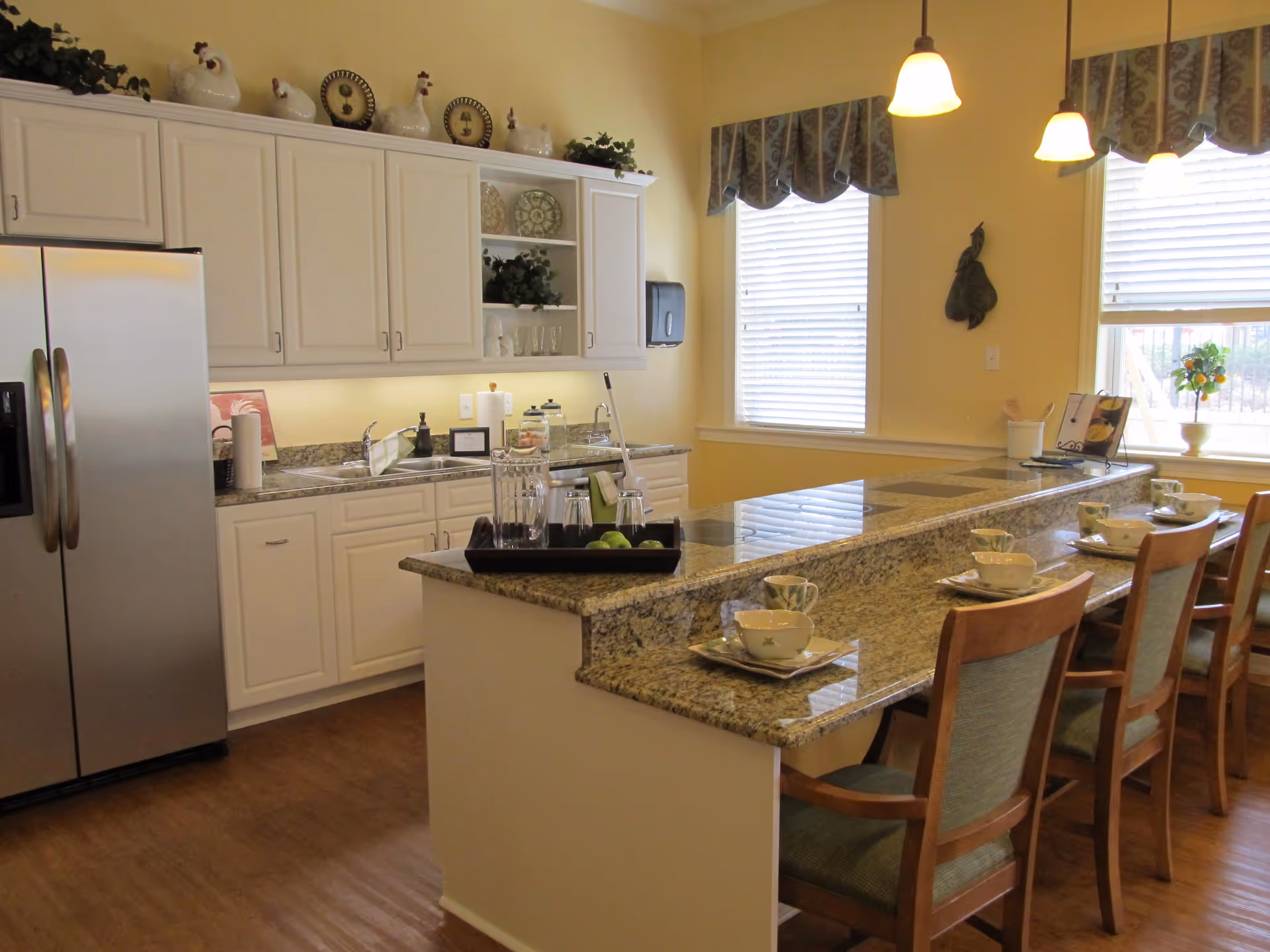 A bright kitchen area with white cabinets, granite countertops, and a stainless steel refrigerator. There is a long kitchen island with four wooden chairs, each set with a cup and saucer. The room has two windows with blinds and decorative valances, and pendant lights hanging from the ceiling. Decorative plates and ceramic chickens are displayed on top of the cabinets.