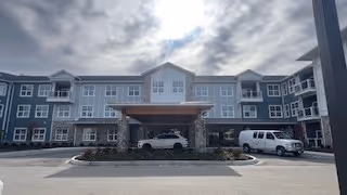 Front exterior view of a large, three-story senior living facility building with multiple windows, a covered entrance supported by stone pillars, and vehicles parked in front under a cloudy sky.