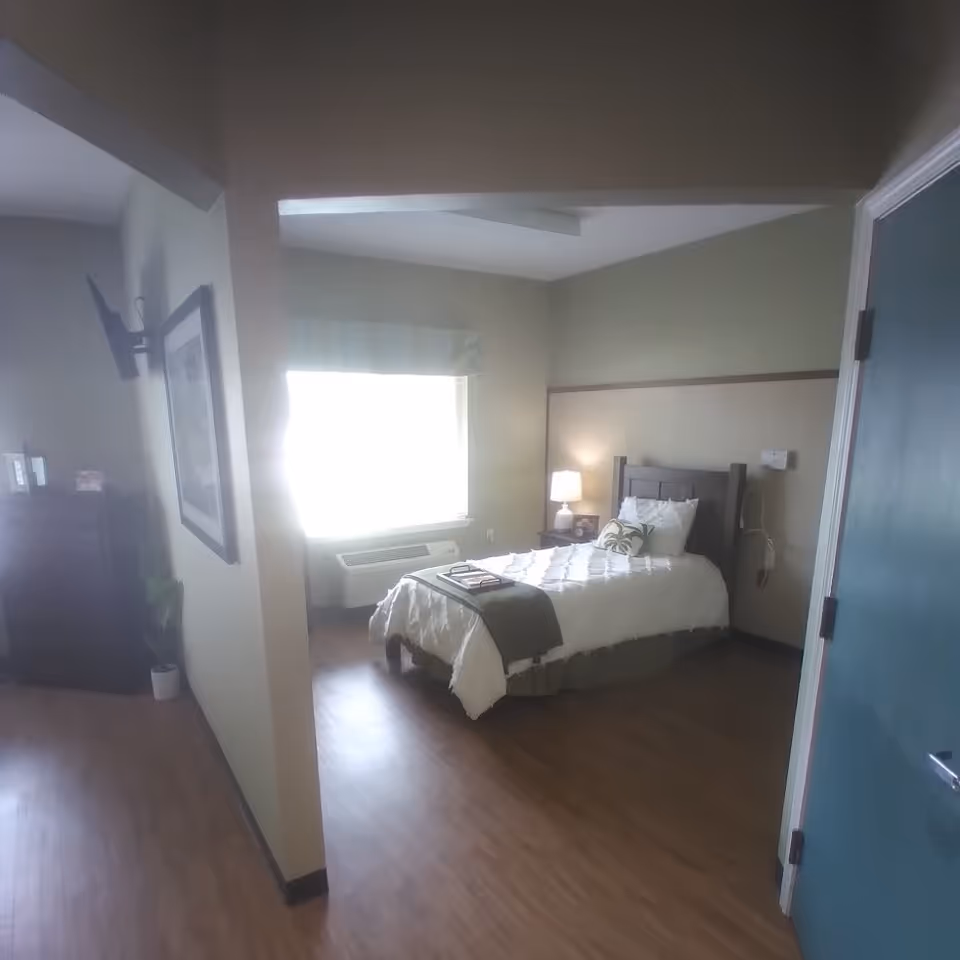 Interior view of a senior living facility bedroom with a single bed made with white bedding and a green throw blanket. A bedside table with a lamp is next to the bed. There is a window with a valance letting in bright light, a wall-mounted TV, and a dresser with some items on top. The floor is wood, and the walls are painted in neutral tones.