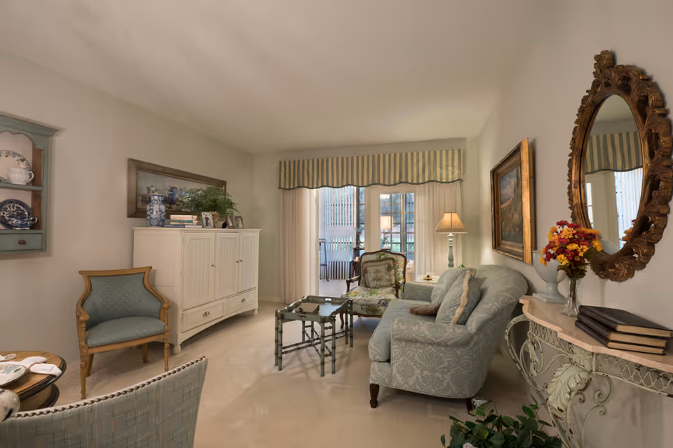 A cozy living room with a light blue patterned sofa, an armchair with floral upholstery, a small green coffee table, and a white cabinet with decorative items on top. There is a large ornate mirror on the wall above a console table with flowers and books. A window with vertical blinds and a striped valance lets in natural light.