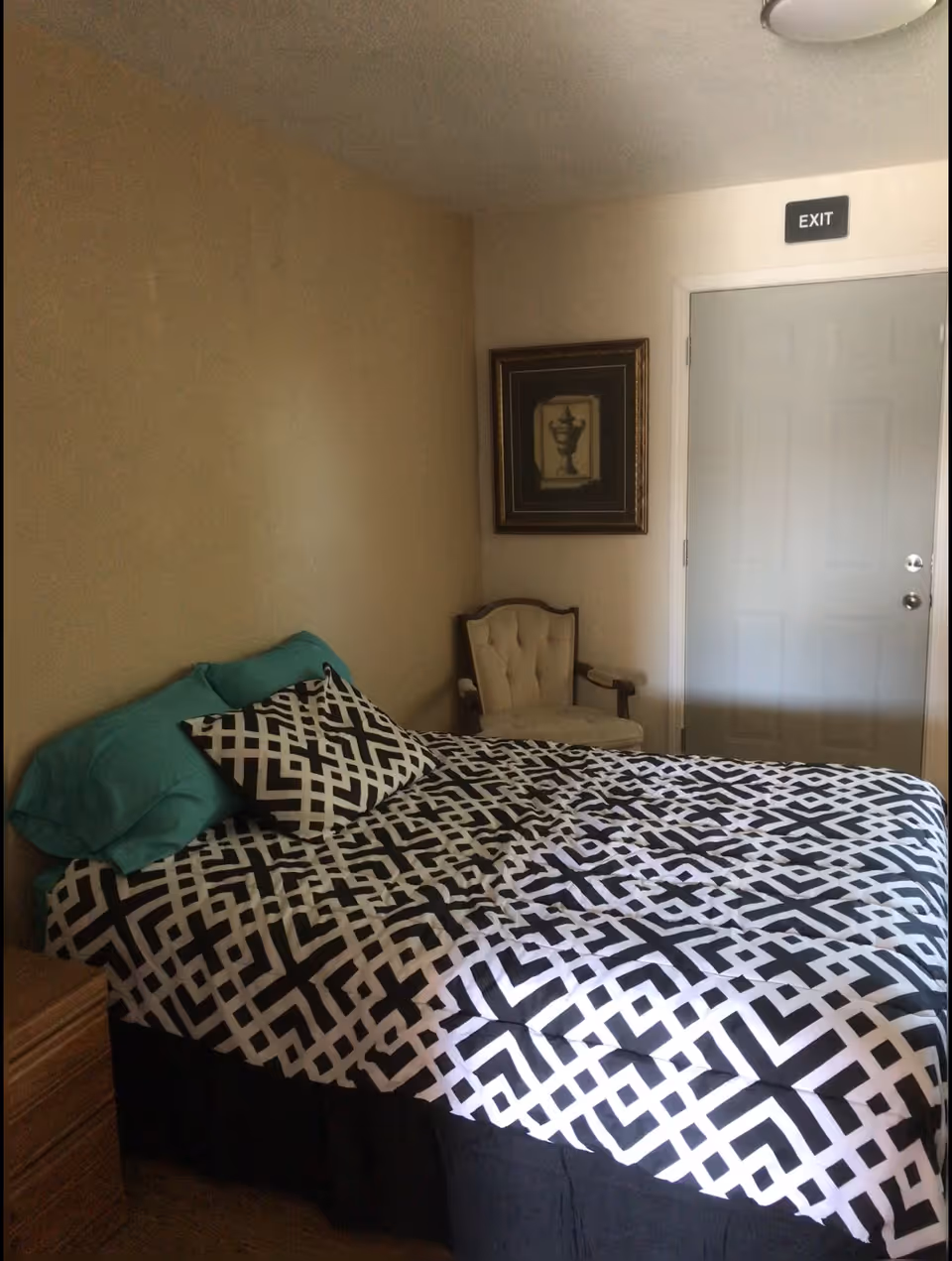 A bedroom with a bed covered in a black and white geometric patterned comforter and two teal pillows. There is a beige upholstered chair in the corner next to a closed door with an 'EXIT' sign above it. A framed picture hangs on the wall above the chair.