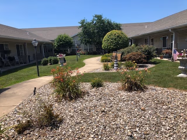 Outdoor courtyard area at Riverbend facility with a paved walkway winding through landscaped garden beds featuring rocks, shrubs, and small trees. Single-story building with covered porch areas and chairs surrounds the courtyard under a clear blue sky.