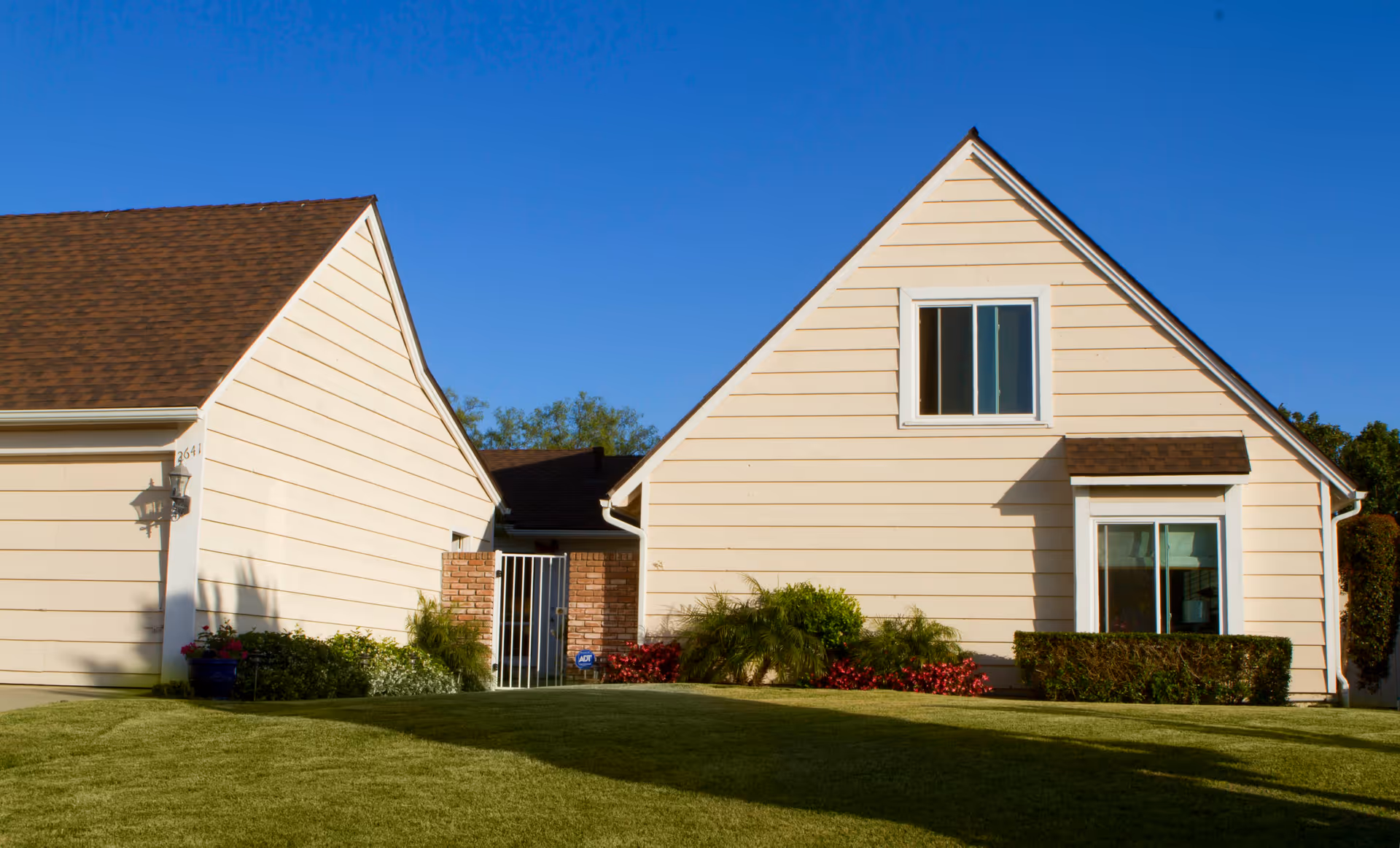 Exterior view of a beige single-story house with a brown shingled roof, two windows, and a well-maintained lawn with shrubs and flowers under a clear blue sky.