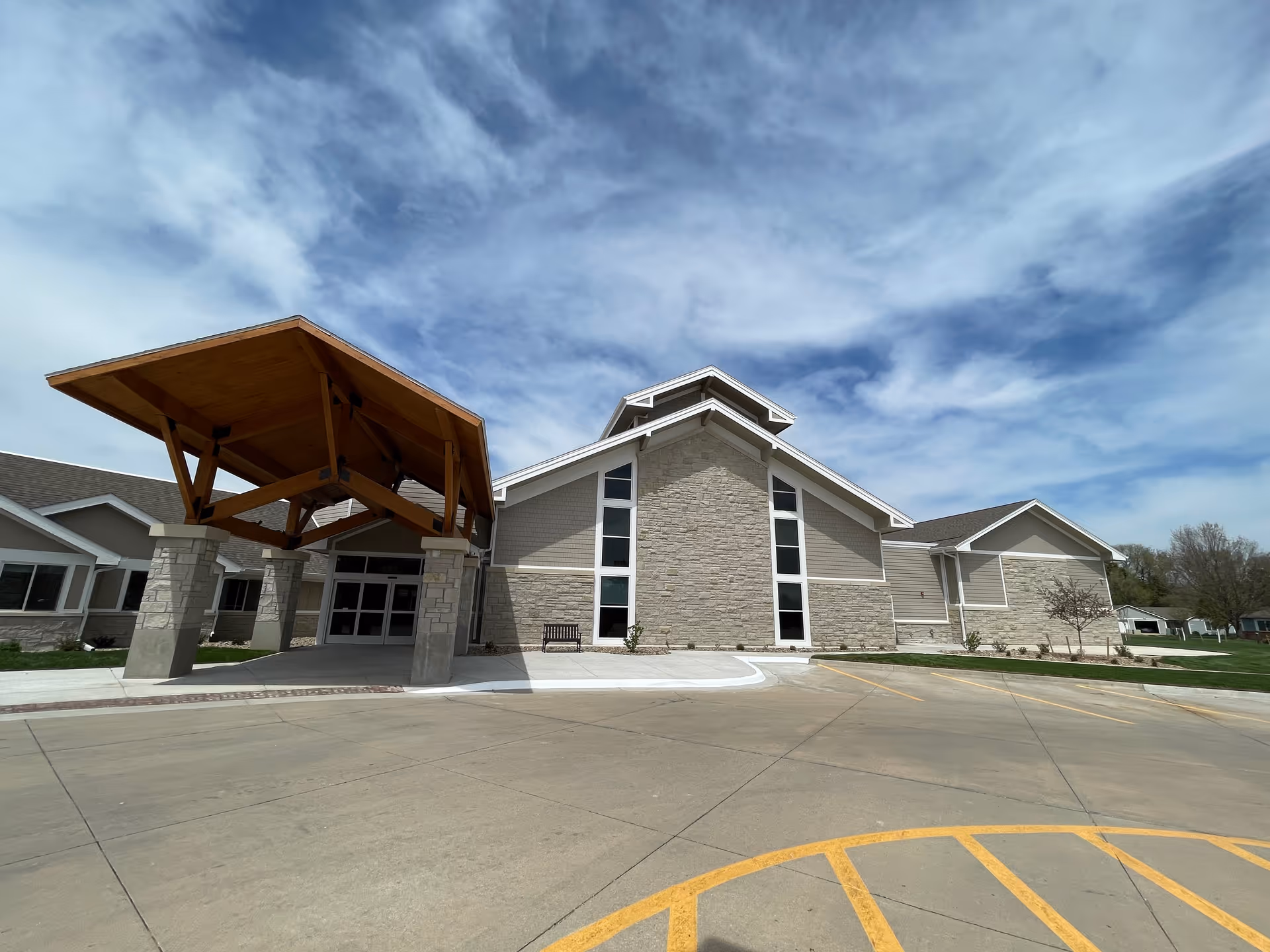 Front entrance of a modern single-story building with a large wooden canopy, stone facade and wide driveway under a cloudy sky.