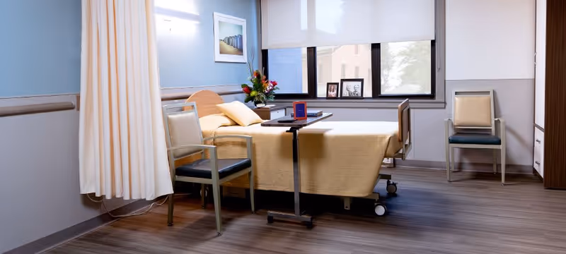 A clean and well-lit hospital or rehabilitation room with a single bed covered in a beige blanket, two chairs, a small overbed table with a book on it, a vase with flowers on a side table, and a window with a view outside partially covered by a roller blind.
