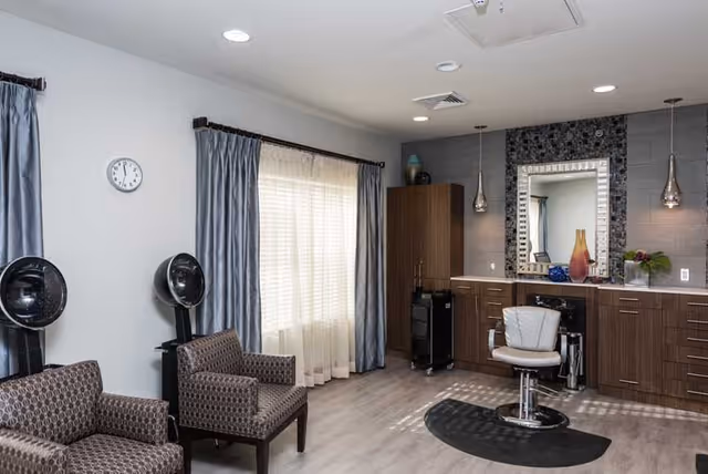 Interior of a salon area in a senior living facility with two patterned armchairs, two black hair dryers on stands, a large window with sheer and blue curtains, a styling chair in front of a counter with cabinets, a large decorative mirror, and two pendant lights hanging from the ceiling.