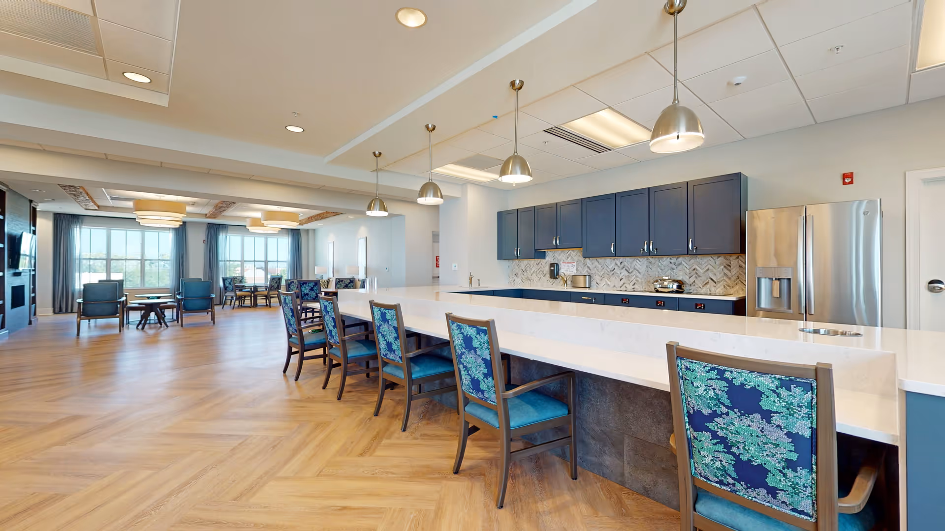 A spacious and modern common area in an assisted living facility featuring a long white countertop with several chairs upholstered in blue and green floral fabric. Behind the counter are blue cabinets, a stainless steel refrigerator, and a tiled backsplash. The room has wood-patterned flooring, pendant lights hanging from the ceiling, and large windows with curtains letting in natural light. In the background, there are additional seating areas with chairs and tables.