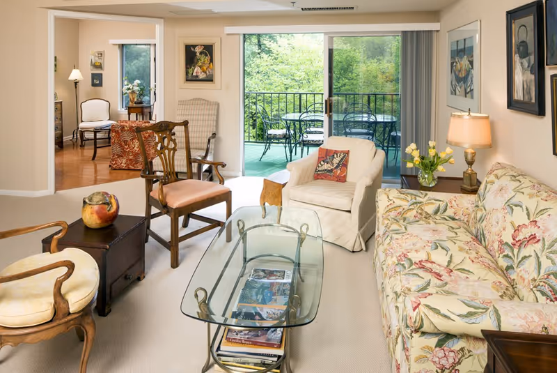 A cozy living room with a floral patterned sofa, a white armchair with a butterfly cushion, and several wooden chairs around a glass coffee table. There is a side table with a lamp and a vase of yellow tulips. The room opens to a balcony with outdoor metal chairs and a table, overlooking green trees. Artwork and framed pictures decorate the walls.