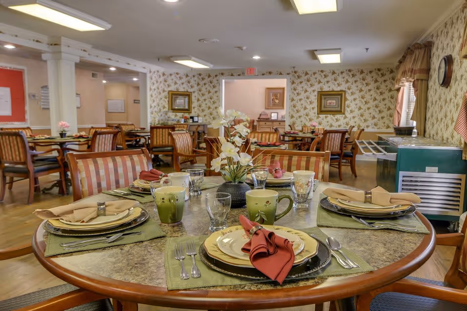 A dining room in a senior living facility with multiple round tables set for meals. Each table has plates, cups, silverware, and cloth napkins, with a floral centerpiece. The room has patterned wallpaper, framed pictures on the walls, and large windows with curtains. Chairs with striped upholstery surround the tables.