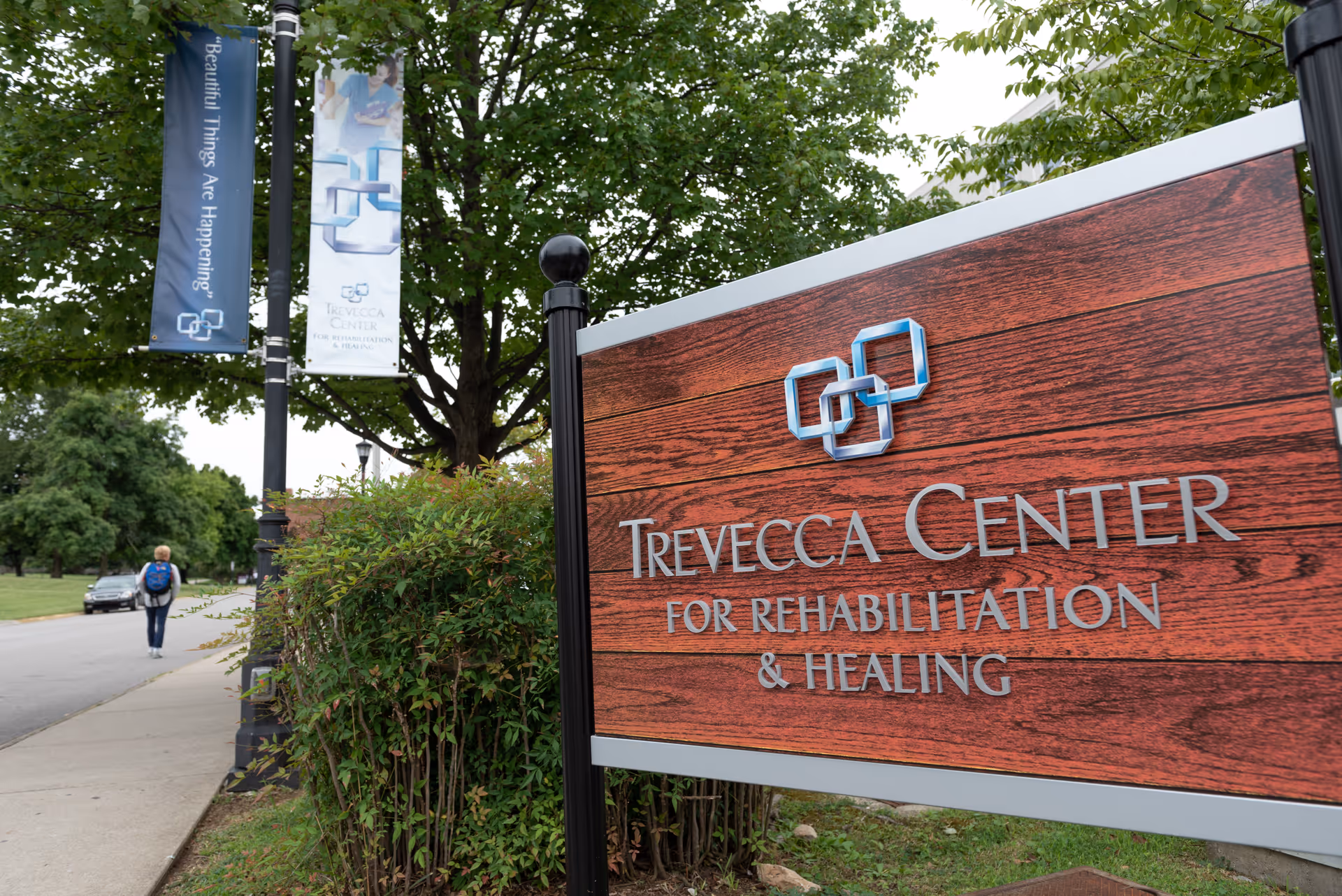 Outdoor view of a wooden sign for Trevecca Center for Rehabilitation & Healing next to a sidewalk with bushes and trees. Two vertical banners hang from a lamp post, one reading 'Beautiful Things Are Happening'. A person with a backpack walks away on the sidewalk.