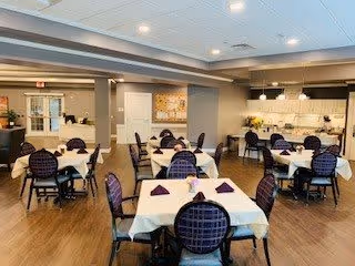 A spacious dining room with multiple tables covered in white tablecloths and set with folded purple napkins. The room features dark wooden chairs with patterned upholstery, wood flooring, and a well-lit kitchen area in the background. The walls are painted in neutral tones, and there is a decorative piece hanging on the far wall.