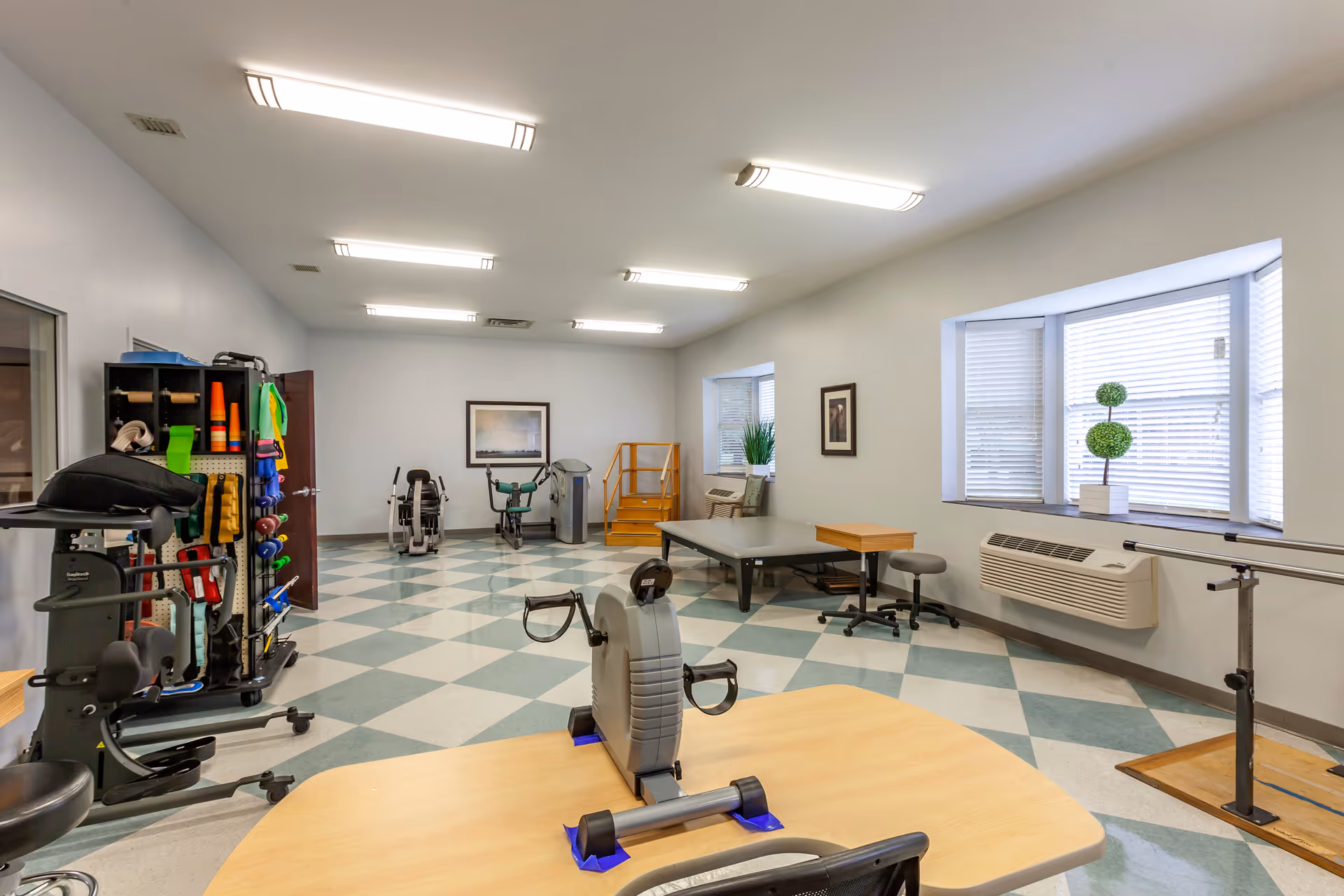 A rehabilitation therapy room with exercise equipment including stationary bikes, hand weights, and therapy tables. The room has checkered flooring, bright overhead lighting, windows with blinds, and framed artwork on the walls.