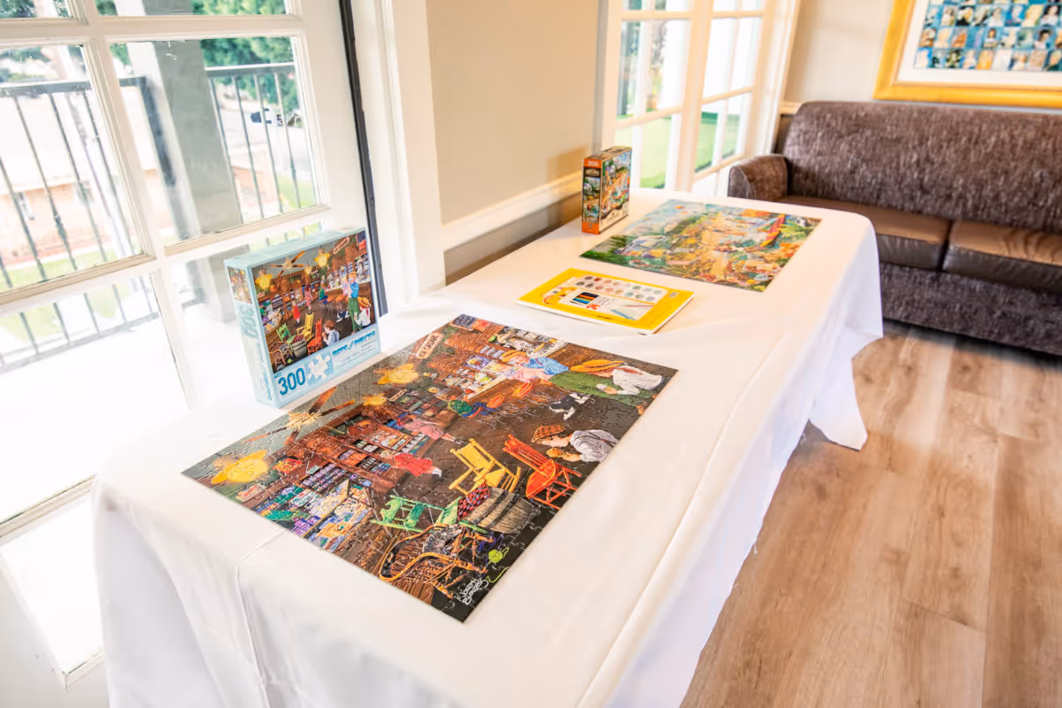 A table covered with a white cloth displaying two completed jigsaw puzzles and their boxes, set near large windows with a view of an outdoor area. In the background, there is a brown couch and a framed picture on the wall.