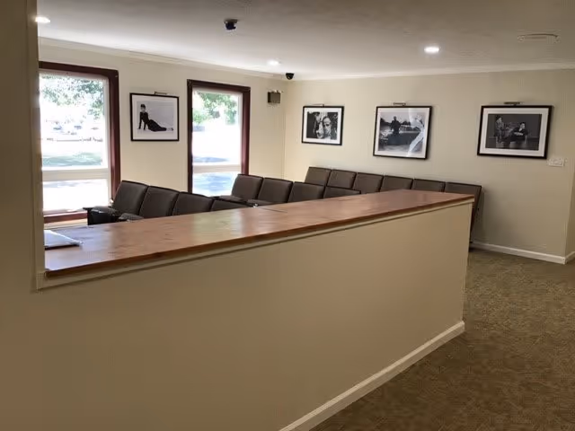 Interior view of a common area with a long wooden counter and several cushioned chairs arranged behind it. The walls are light-colored and decorated with framed black and white photographs. Large windows allow natural light to enter the room.