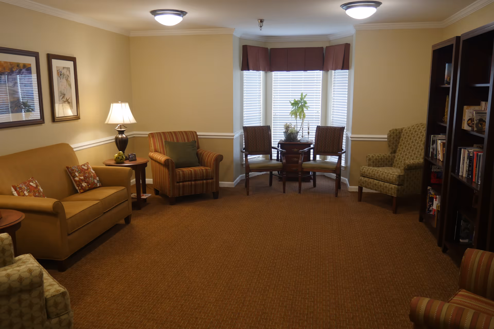 A cozy living room with beige walls and brown carpet. The room features a mustard yellow sofa with patterned cushions, a striped armchair with a green pillow, two wooden chairs with green cushions around a small round table by a window with white blinds and a brown valance. There is a patterned armchair near a dark wooden bookshelf filled with books. A table lamp with a white shade sits on a side table next to the sofa. Two framed pictures hang on the wall above the sofa.