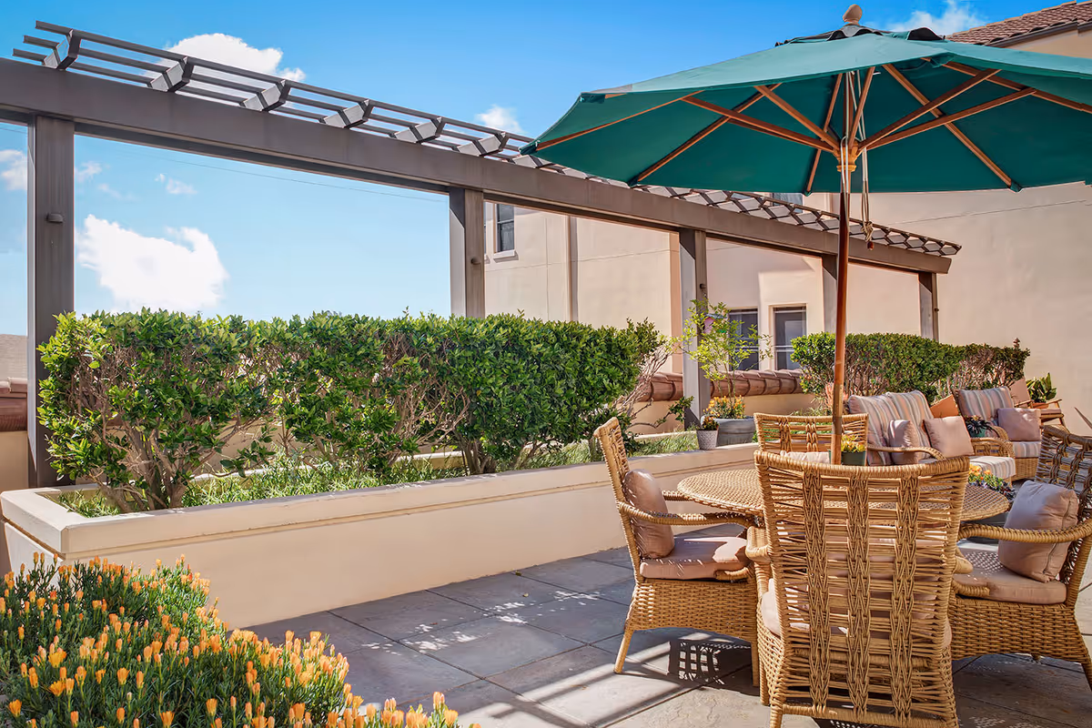 Outdoor patio with a wicker dining set under a green umbrella, surrounded by hedges and a pergola.