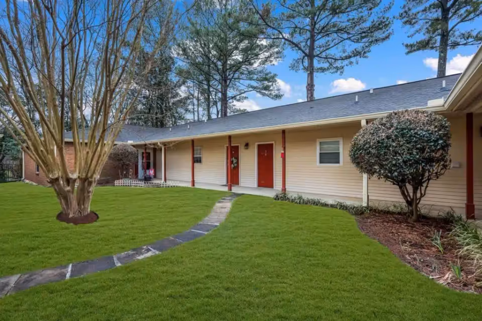 Exterior view of a single-story building with beige siding and red doors, surrounded by a well-maintained lawn, a stone pathway, and trees under a blue sky with some clouds.