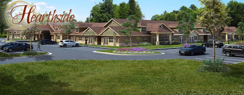 Exterior view of Hearthside Senior Living facility showing a single-story building with multiple peaked roofs, surrounded by a parking lot with several cars and landscaped greenery including trees and flower beds under a clear blue sky.