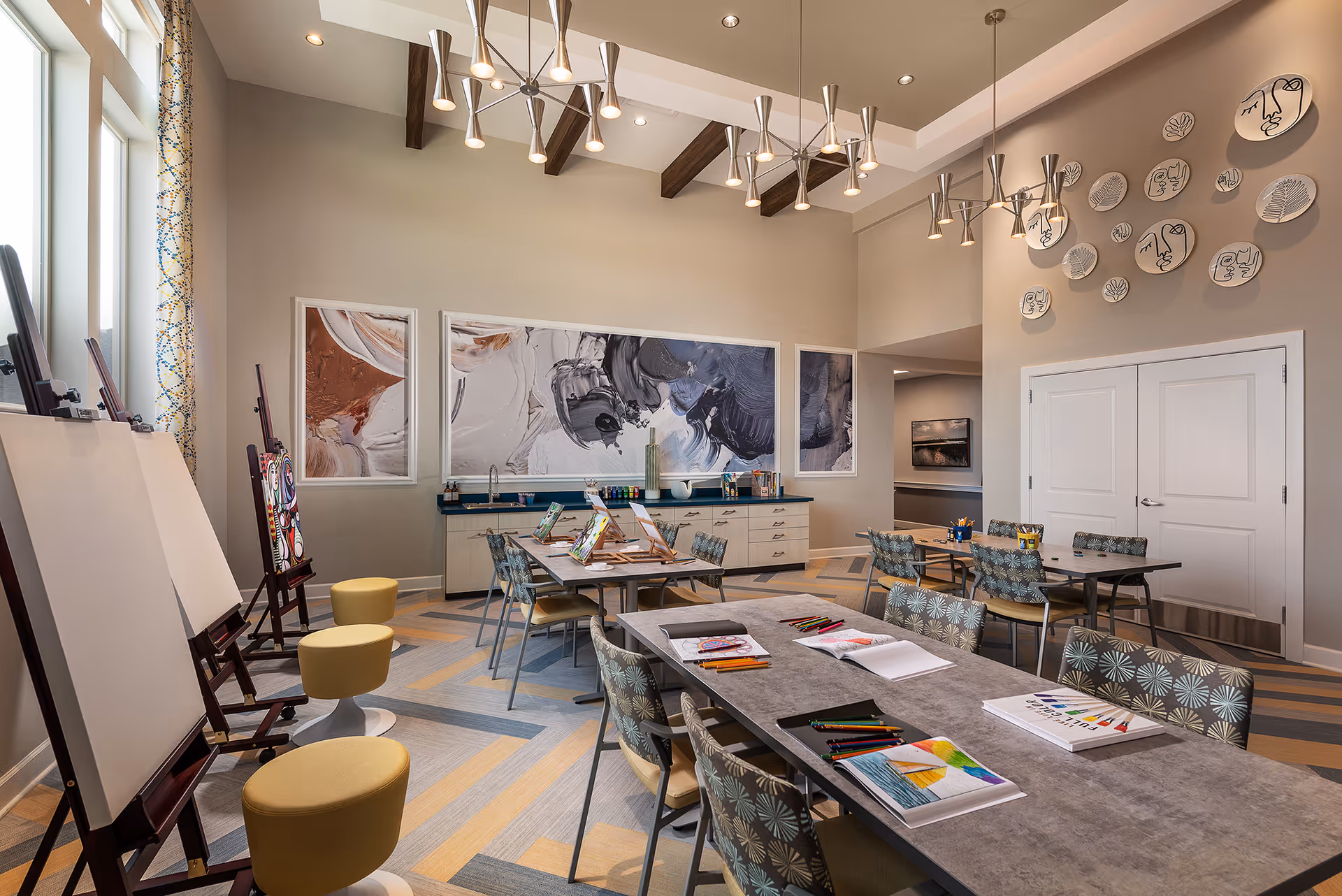 A bright and spacious art room with several tables and chairs arranged for painting and drawing activities. There are easels with blank canvases, colorful stools, and art supplies like colored pencils and paintbrushes on the tables. The walls are decorated with abstract art and artistic plates featuring face designs. The room has large windows allowing natural light to fill the space, and modern pendant lights hang from the ceiling.