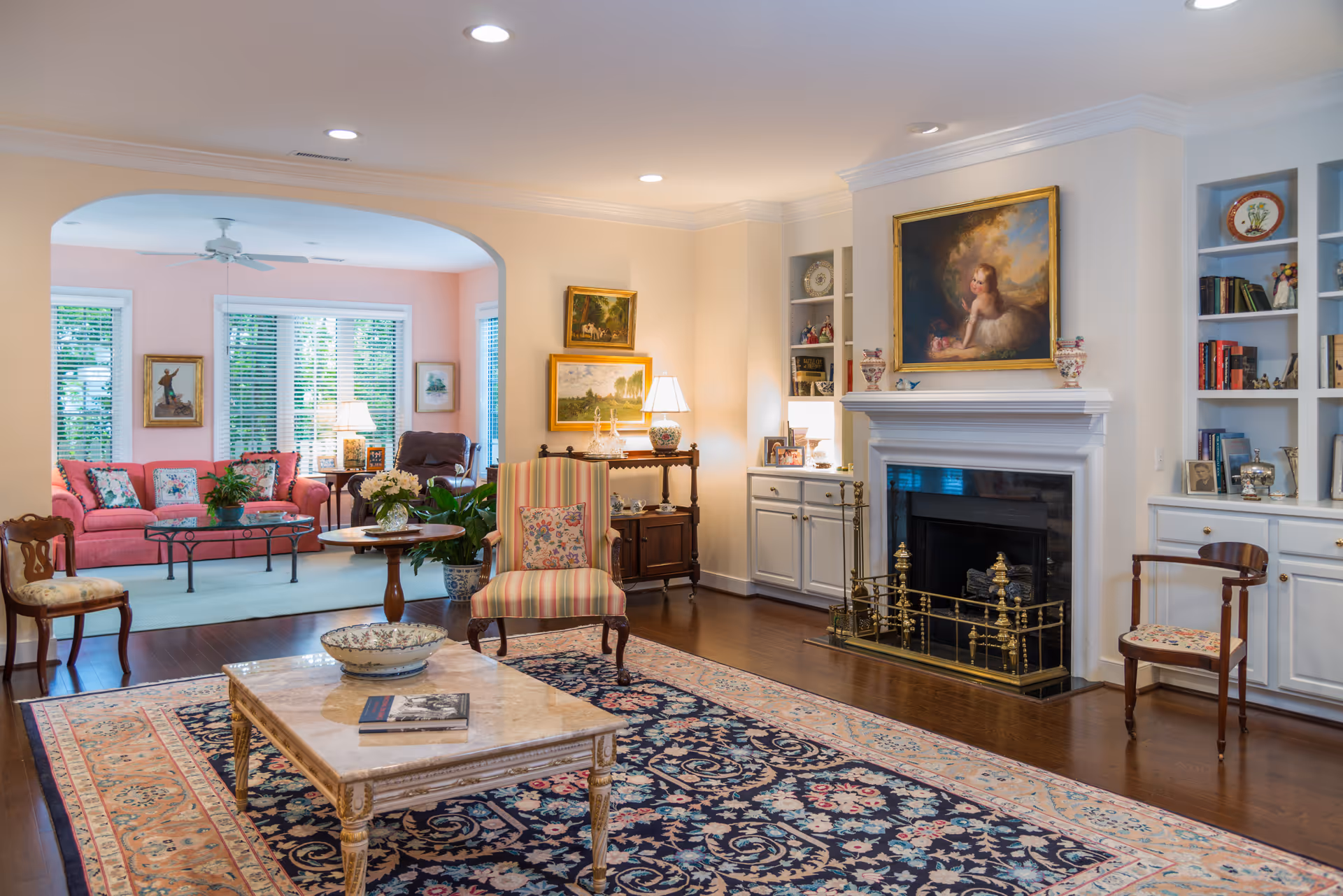 Well-appointed living room with a fireplace, built-in bookshelves, patterned rug, coffee table and multiple seating areas opening to a sunlit sitting room with a coral sofa.