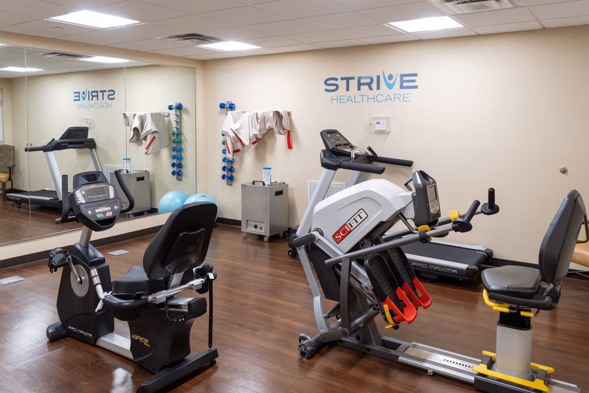 Indoor fitness room with exercise equipment including a recumbent bike, a treadmill, and a multi-exercise machine. The room has wooden flooring, a large wall mirror, and a wall-mounted rack with towels and dumbbells. The wall features the logo 'STRIVE HEALTHCARE'.