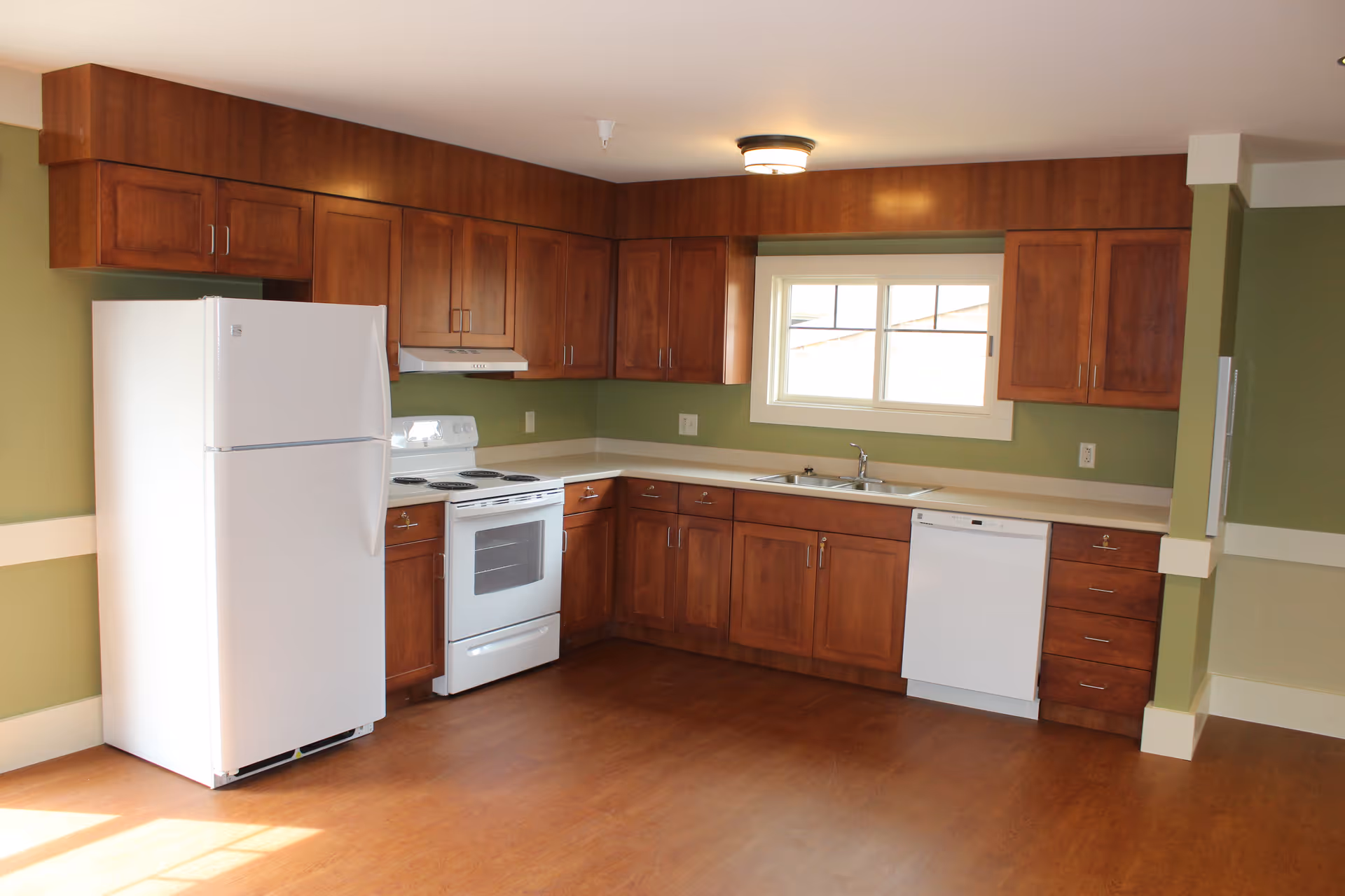 A kitchen with wooden cabinets and green walls featuring a white refrigerator, white stove with oven, white dishwasher, and a double sink under a window. The floor is wooden and the room is well-lit with natural light.