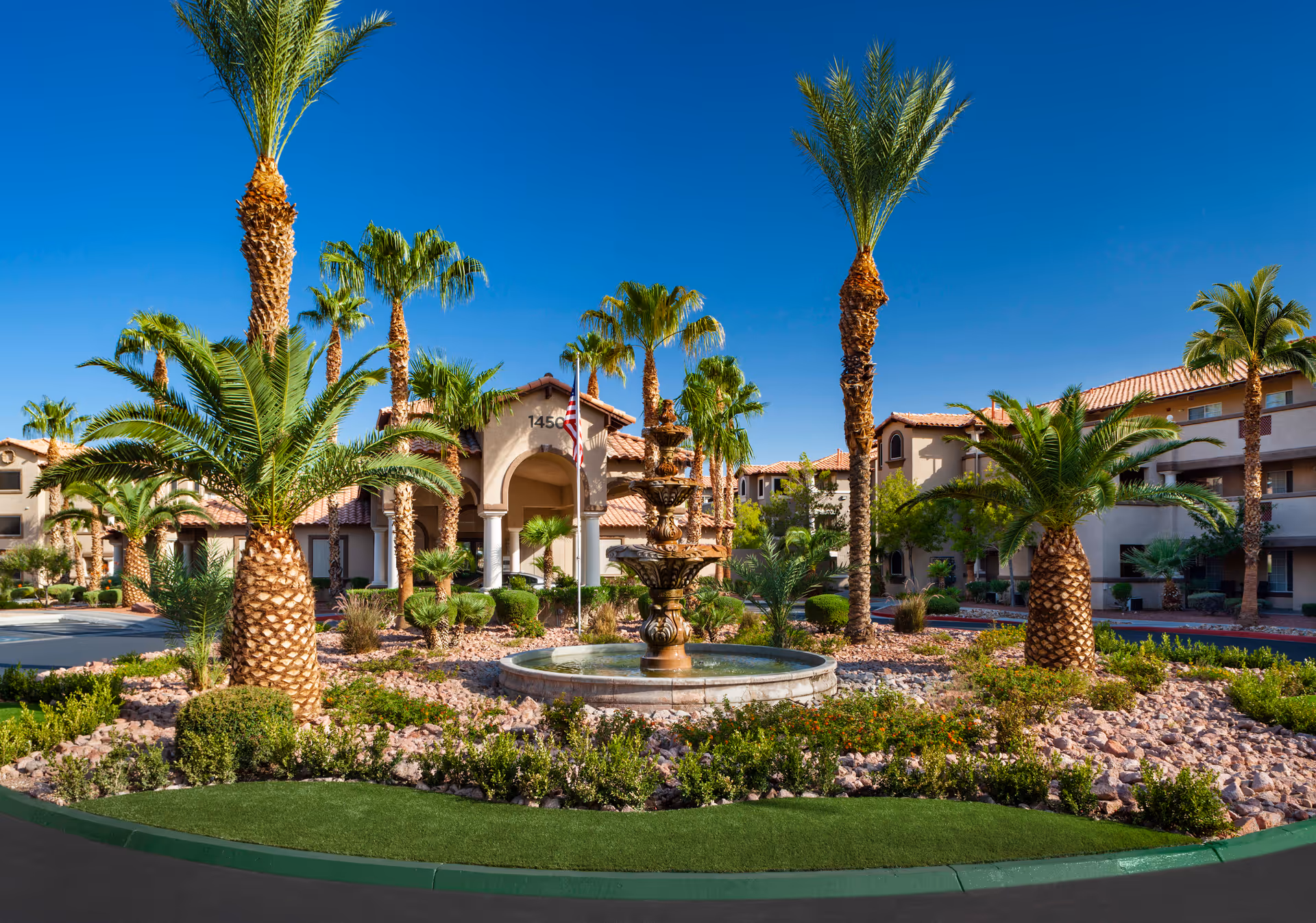 Exterior view of Destinations Pebble senior living facility featuring a landscaped roundabout with a tiered water fountain in the center, surrounded by palm trees and shrubs. The building has a beige stucco finish with red tile roofing under a clear blue sky.