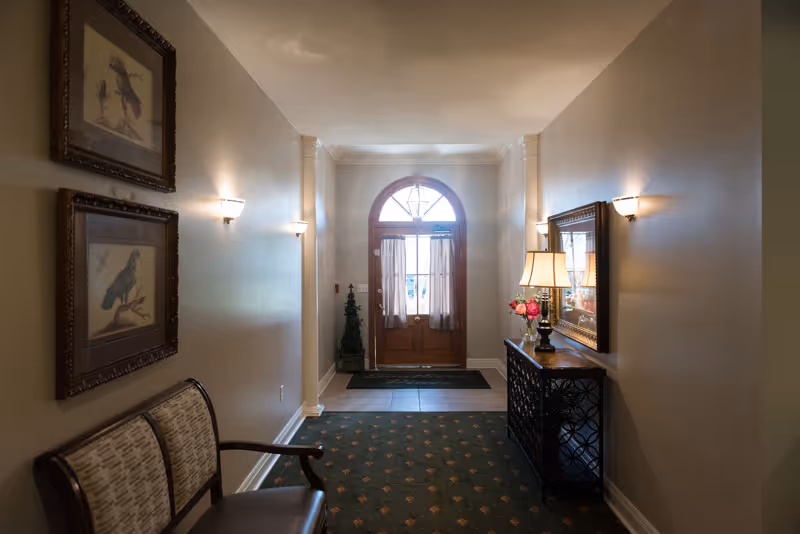 A hallway with beige walls and a patterned green carpet leading to a wooden door with a window and curtains. On the left wall, there are two framed bird pictures above a cushioned bench. On the right side, there is a console table with a lamp, flowers, and a large mirror above it. Wall sconces provide lighting along the hallway.