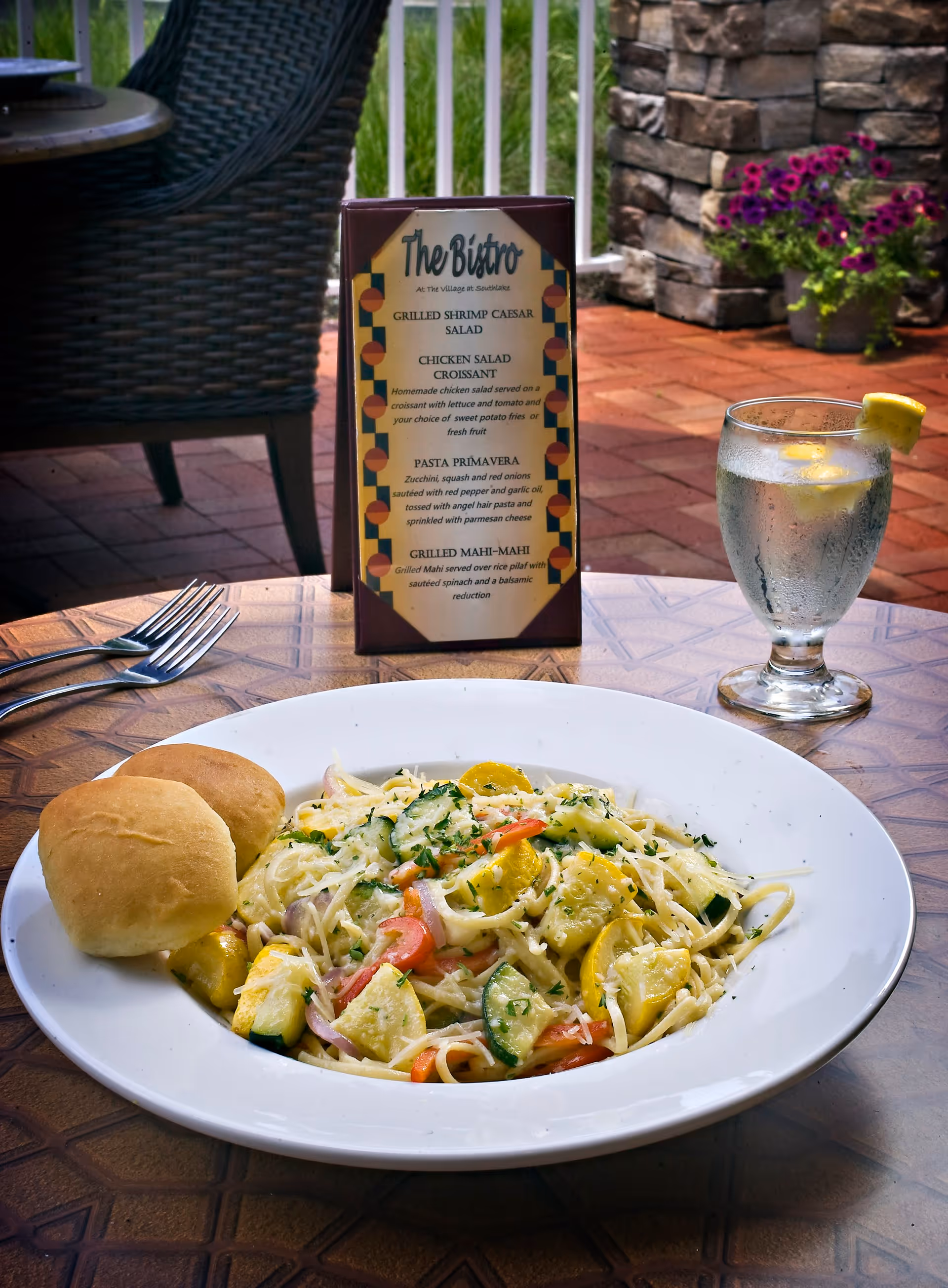 A plate of pasta primavera with zucchini, squash, red onions, and red peppers, served with two bread rolls on a wooden table. A glass of water with a lemon wedge is placed to the right. Behind the plate is a menu titled 'The Bistro at The Village at Southlake' listing various dishes. The setting appears to be an outdoor patio with a wicker chair, stone wall, and potted flowers in the background.