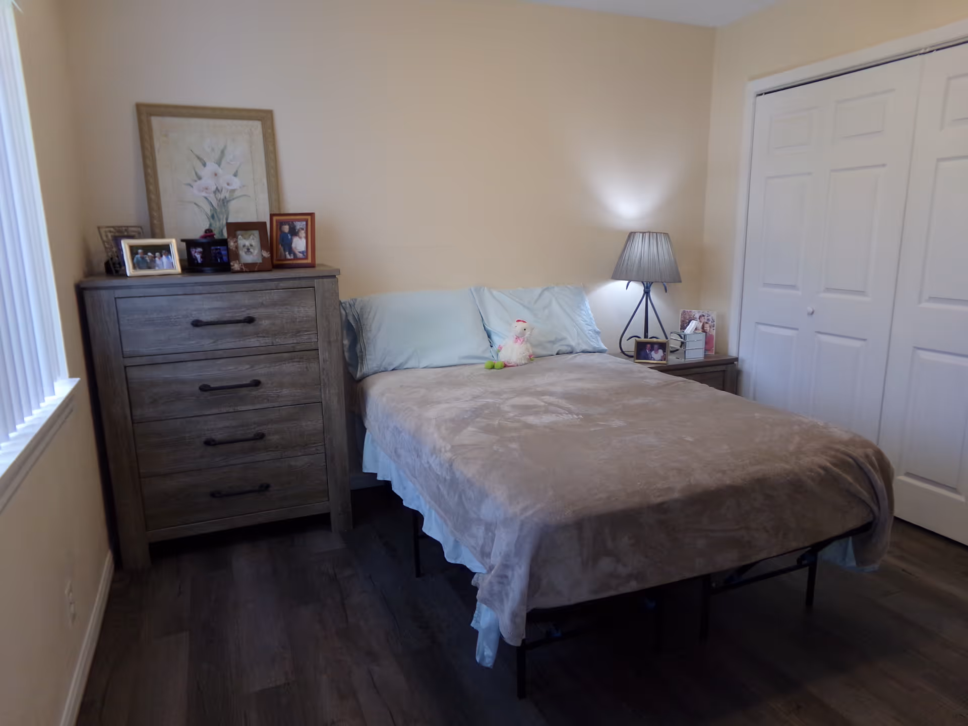 Small bedroom with a bed covered by a beige blanket, a wooden dresser topped with framed photos, a nightstand with a lamp, and closet doors.