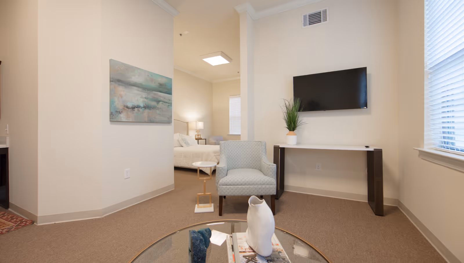 A bright sitting area with an armchair, wall-mounted TV, console table and a glimpse of a bed in the adjoining bedroom.