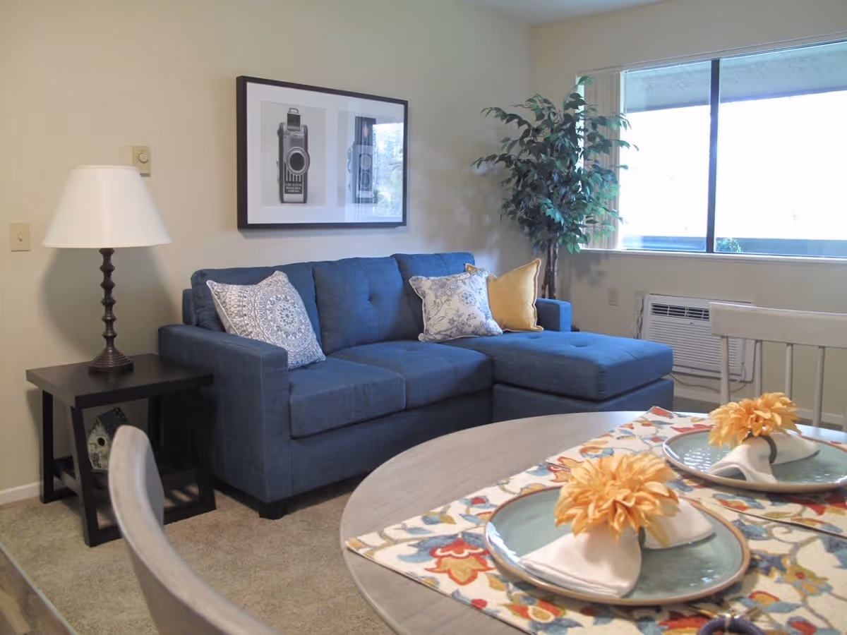 Cozy living room area with a blue sectional sofa adorned with three decorative pillows. A black side table with a lamp is positioned next to the sofa. A framed black and white photo of a vintage camera hangs on the wall above the sofa. A large window with vertical blinds lets in natural light, and a green potted plant is placed near the window. In the foreground, a round dining table is set with two plates, each with a folded white napkin topped with a yellow flower, on a colorful patterned table runner.