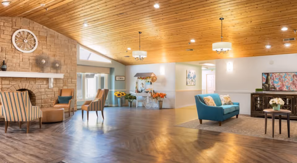 Open, well-lit senior living lounge with a wood-paneled ceiling, stone fireplace, multiple seating areas, and decorative plants.
