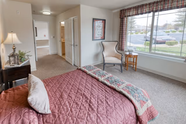 A bright senior living bedroom with a large window overlooking a parking lot. The room features a bed with a maroon quilt and a decorative pillow, a wooden nightstand with a lamp and plant, a comfortable armchair next to a small round wooden table, and a framed picture on the wall. The room has beige walls and carpeted floor, with an open doorway leading to another room and a closet with double doors.