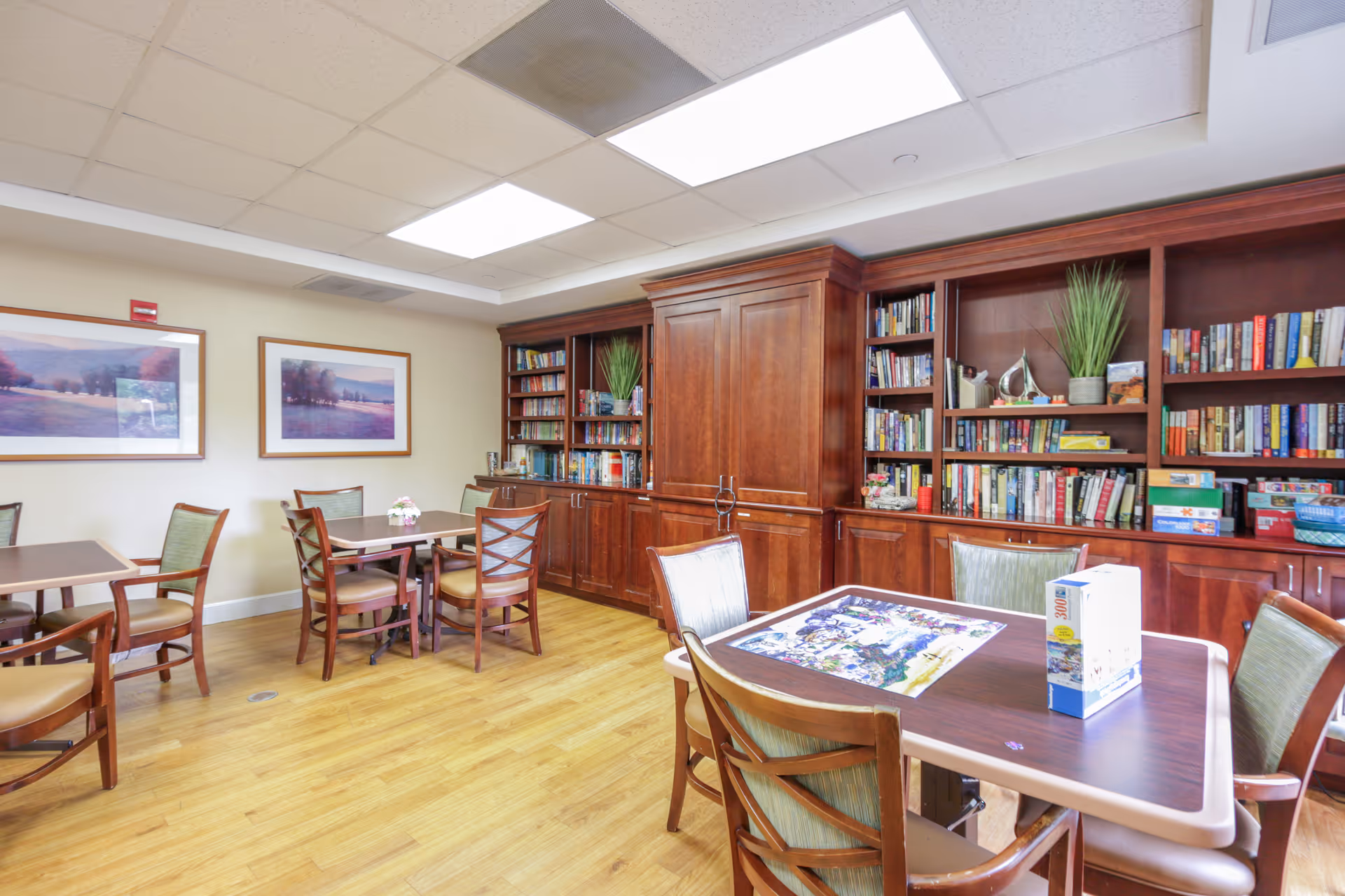 A well-lit room with wooden flooring and several tables surrounded by chairs. One table has a partially completed jigsaw puzzle and a puzzle box on it. The back wall features large wooden bookshelves filled with books, plants, and board games. Two framed landscape paintings hang on the left wall.