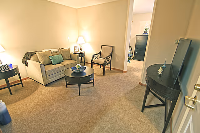 A cozy living room area with a beige loveseat, a wooden chair, a round coffee table with a small flower arrangement, and two side tables with lamps. The room has beige carpet and walls, and an open doorway leads to another room with a dresser and a plant.