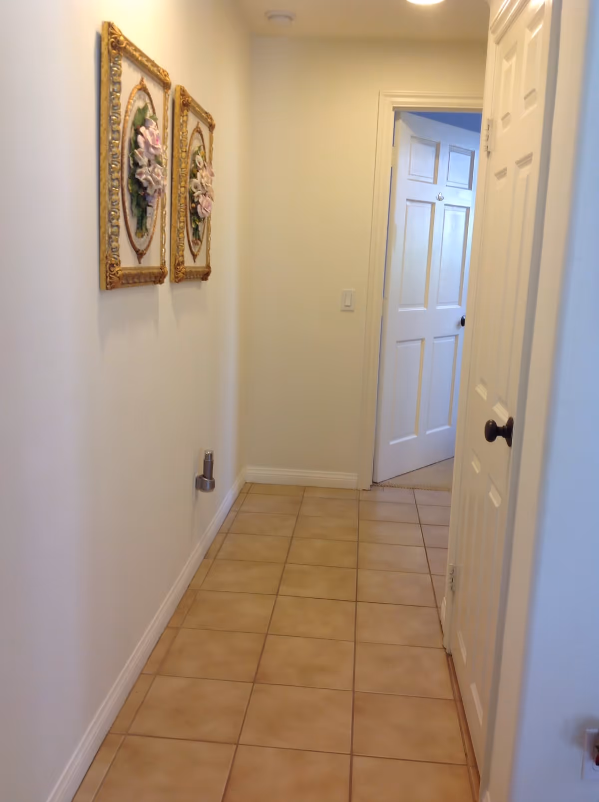 A narrow hallway with beige tiled floor and white walls. Two decorative framed floral artworks hang on the left wall. At the end of the hallway, there is an open white door leading to another room with carpeted flooring. Another white door is partially visible on the right side of the hallway.
