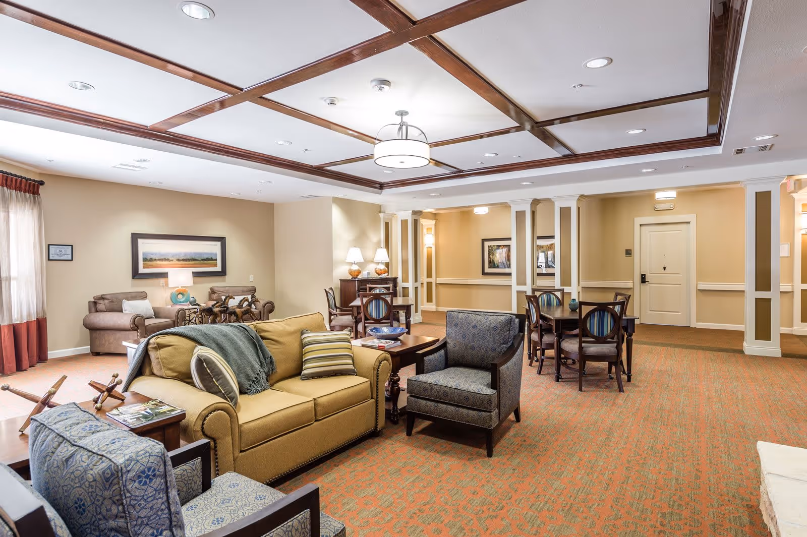 A spacious and well-lit living room area in a senior living facility with beige walls and a patterned orange carpet. The room features a yellow sofa with pillows and a throw blanket, two patterned armchairs, a wooden coffee table, and a side table with magazines. In the background, there are additional seating areas with armchairs, tables, lamps, and framed artwork on the walls. The ceiling has wooden beams and recessed lighting.
