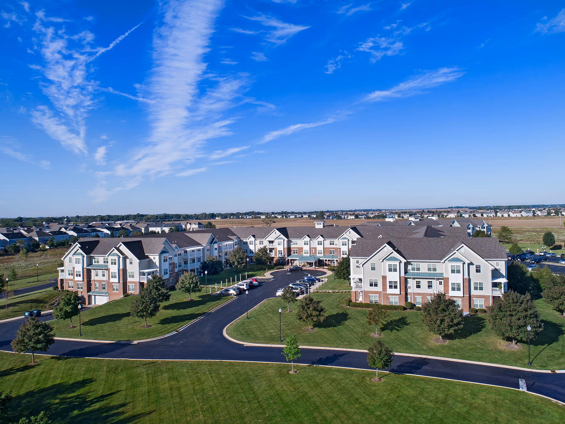 Aerial view of a large senior living facility named American House Cedarlake, featuring multiple connected buildings with beige and brick exteriors, surrounded by well-maintained green lawns, trees, and a curved driveway with parked cars under a bright blue sky with wispy clouds.